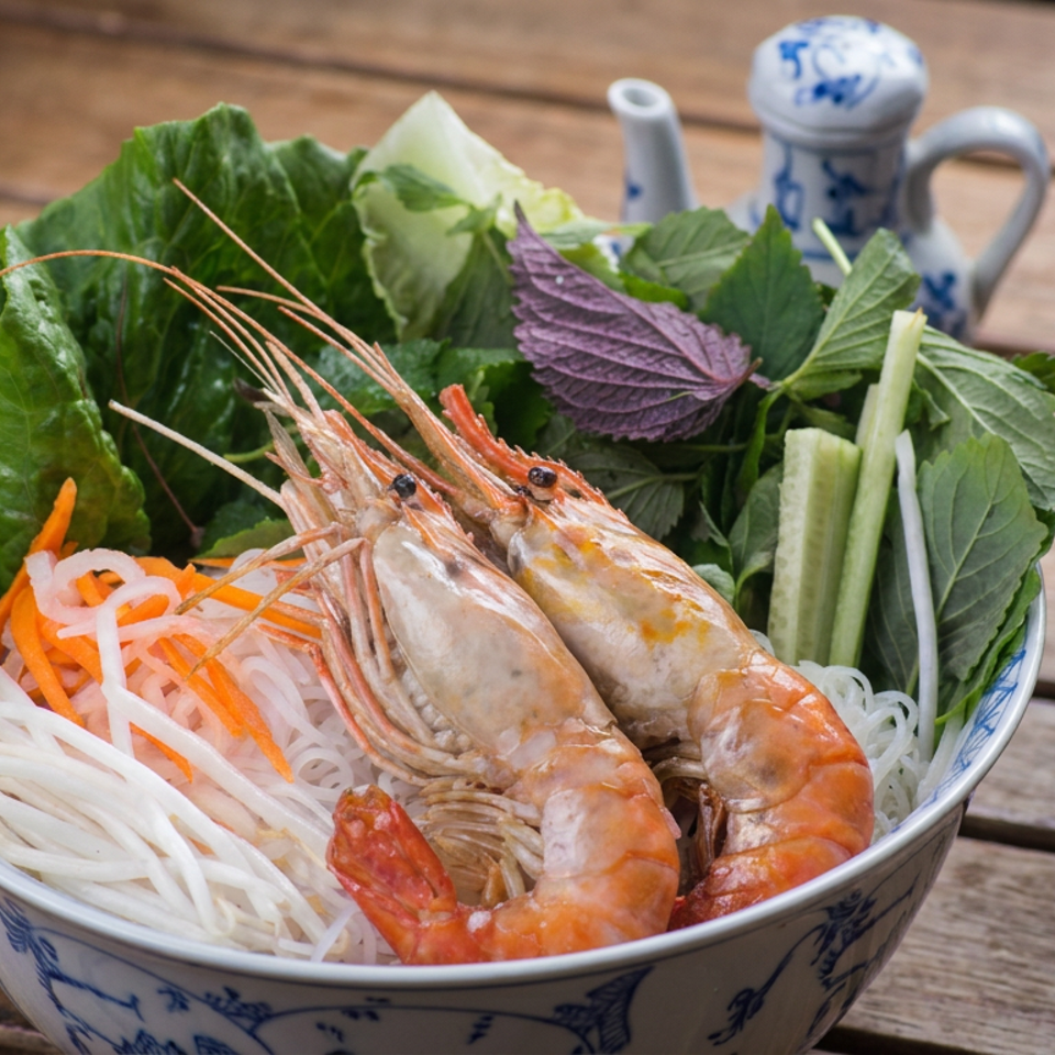 Vietnamese noodle bowl topped with prawns, lettuce, herbs, cucumber, carrots, and bean sprouts beside dipping sauce and beer.