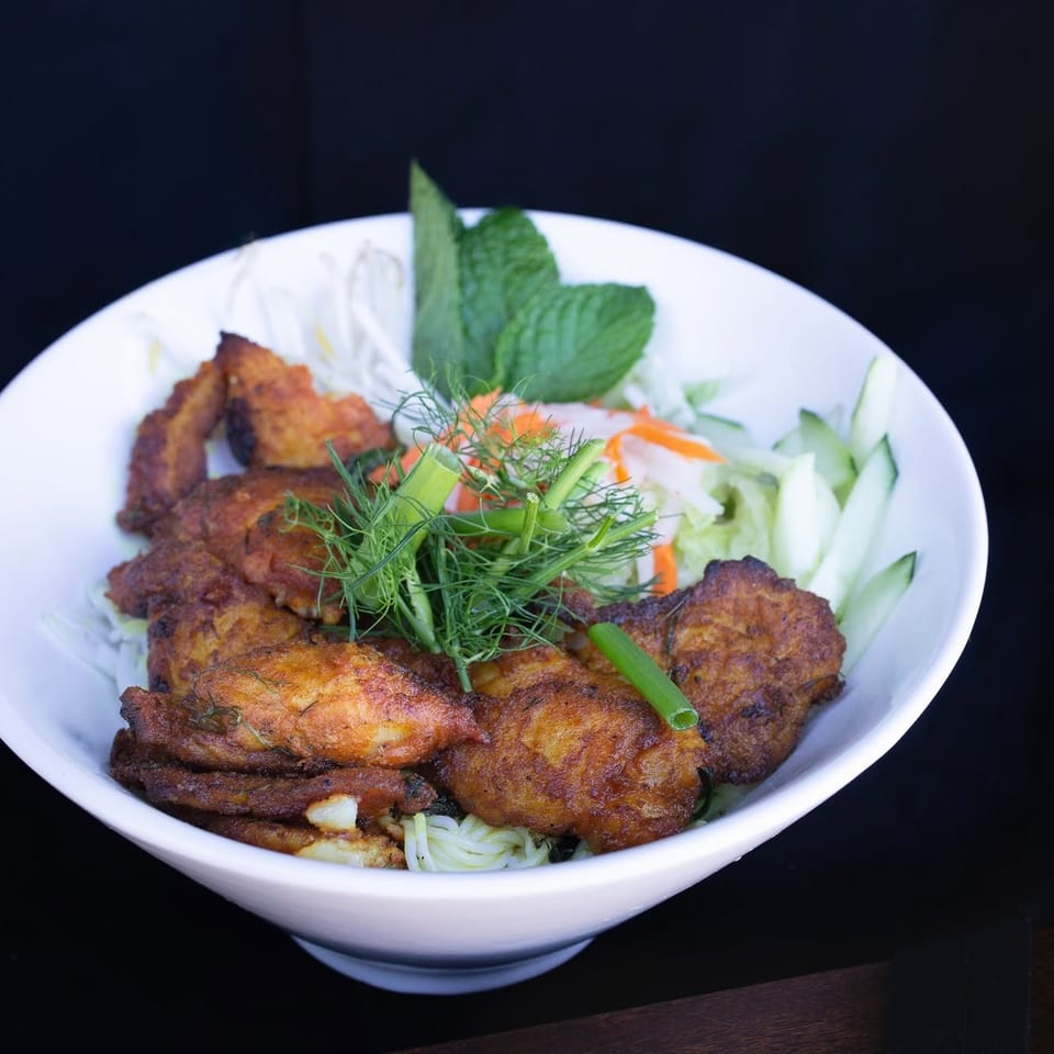 Bowl of crispy golden fritters over noodles and vegetables, garnished with mint, dill, carrots, cucumber, and green onion.
