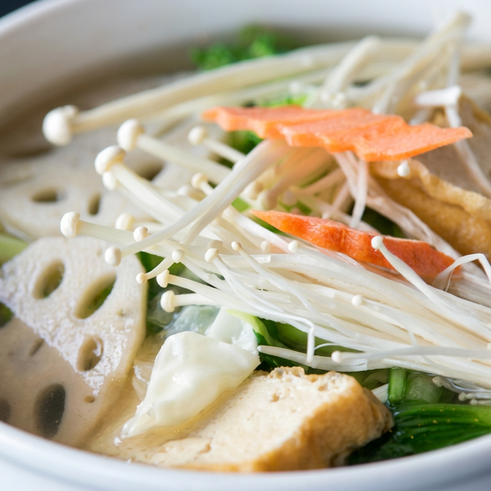 A bowl of Asian vegetable soup topped with enoki mushrooms, tofu, lotus root, carrots, greens, and a dumpling.