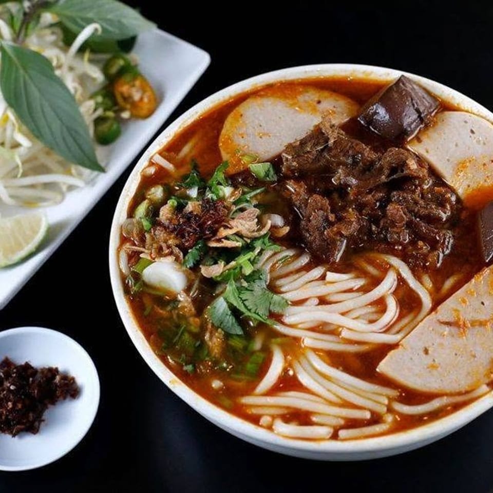 Bowl of spicy Vietnamese noodle soup with beef, pork sausage, herbs, eggplant, bean sprouts, lime, and chili paste.