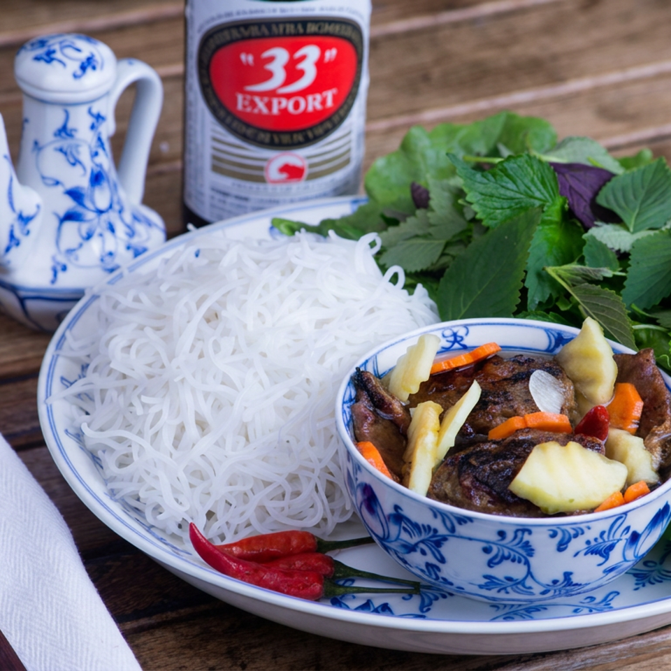 Vietnamese rice noodles, grilled meat, pickled vegetables, fresh herbs, and chilies arranged on a blue-and-white plate beside beer and teapot.